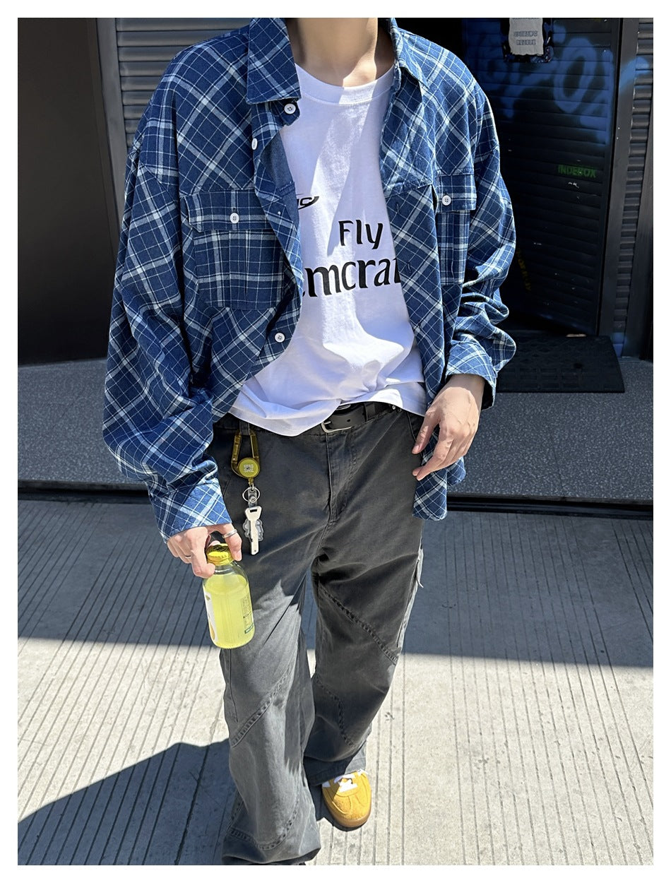 Person wearing the blue plaid shirt unbuttoned over a white graphic tee and gray pants, cropped view holding a drink