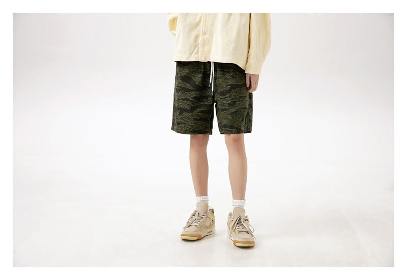 Front three-quarter view of a model wearing dark green camouflage shorts with a cream top and tan sneakers.