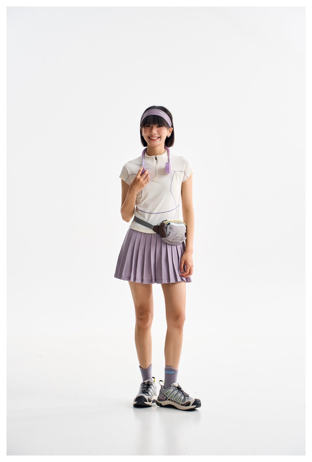 Model wearing a white short-sleeve zip top and pleated lilac skirt, with a lilac headband and waist bag.