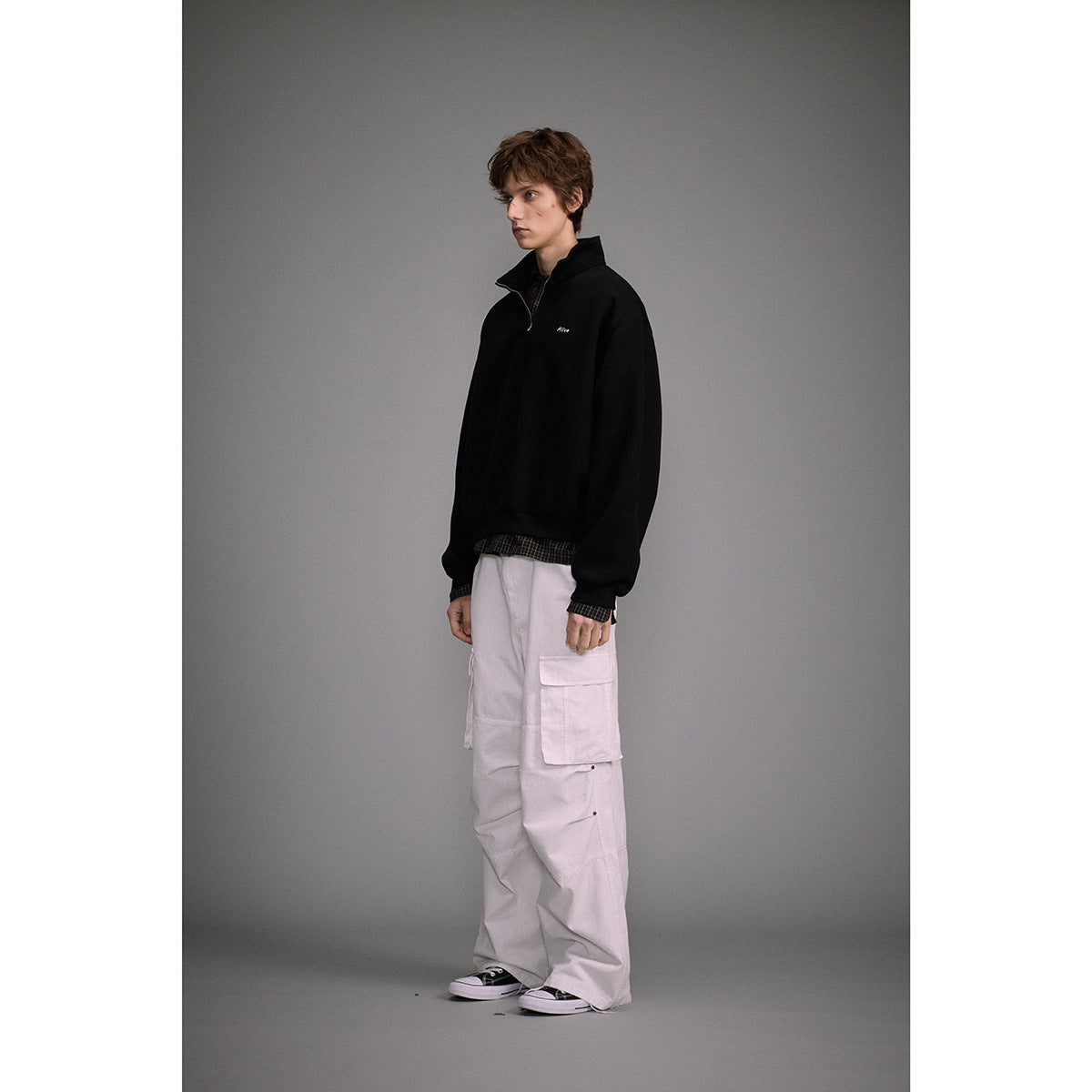 Full-length model wearing a black quarter-zip sweatshirt with white chest embroidery and loose light cargo pants.