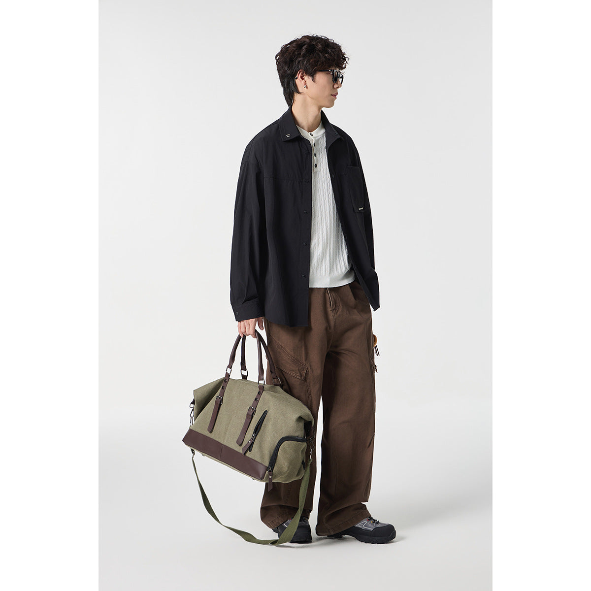 Model wearing a black oversized button-front shirt over a white knit top, with brown trousers and a duffel bag.