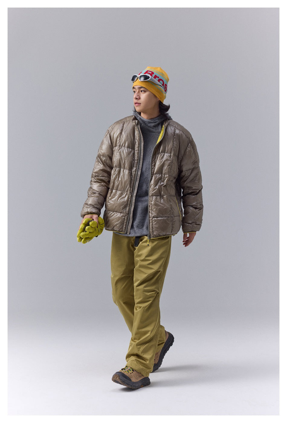 Model walking in a taupe quilted jacket over a gray top with olive pants and a yellow beanie.