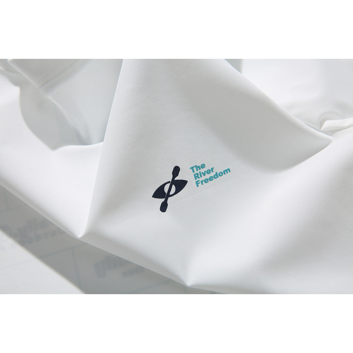 Close-up of a dark paddle emblem and turquoise “The River Freedom” text printed on white T-shirt fabric.