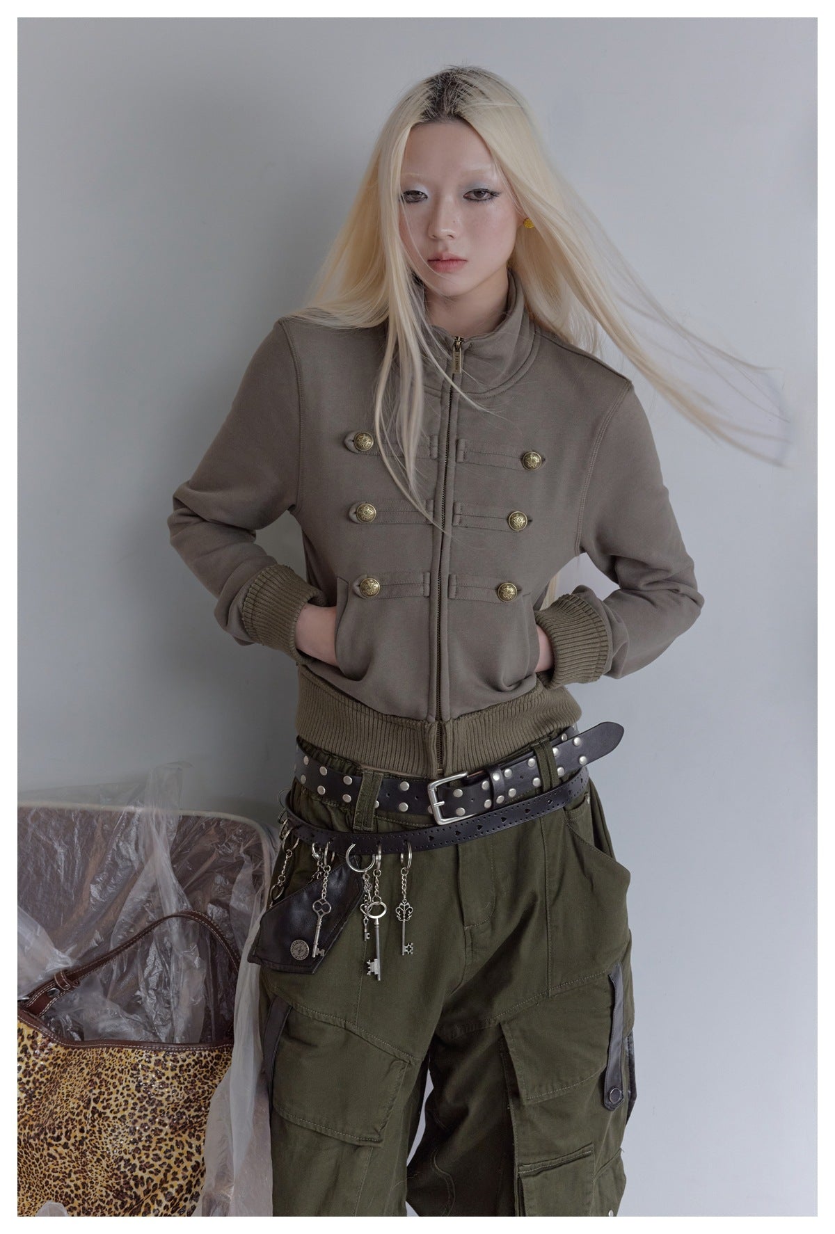 Model wearing a taupe cropped zip jacket with brass button tabs, ribbed cuffs, and an olive waistband below.