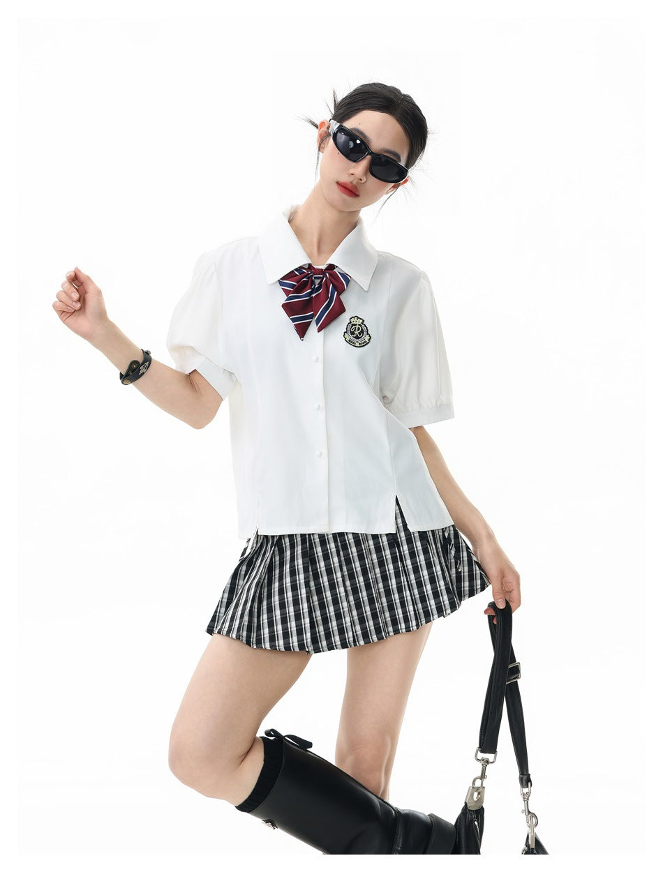 Model in a white puff-sleeve shirt with a striped bow and crest patch, posed with a pleated plaid mini skirt and boots
