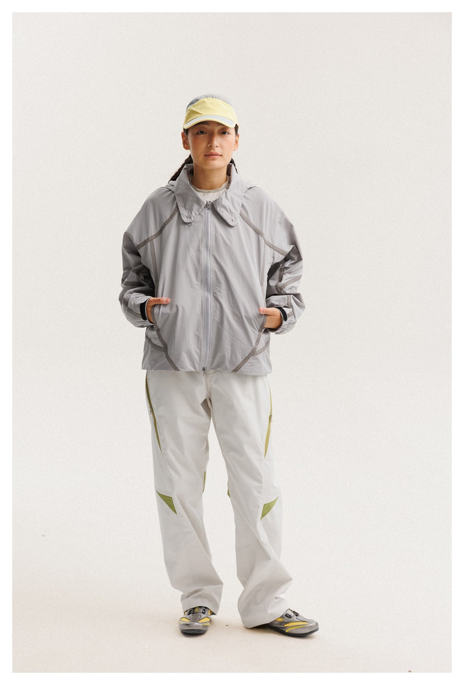 Full-length model wearing a light gray zip-front hooded jacket with dark seam details and white pants with olive accents.
