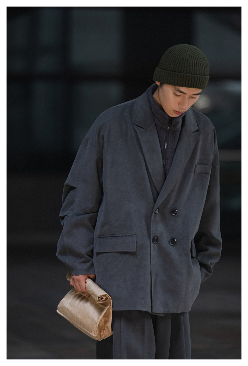 Model wearing a slate-gray double-breasted coat over a dark zip-neck top, with an olive ribbed beanie.