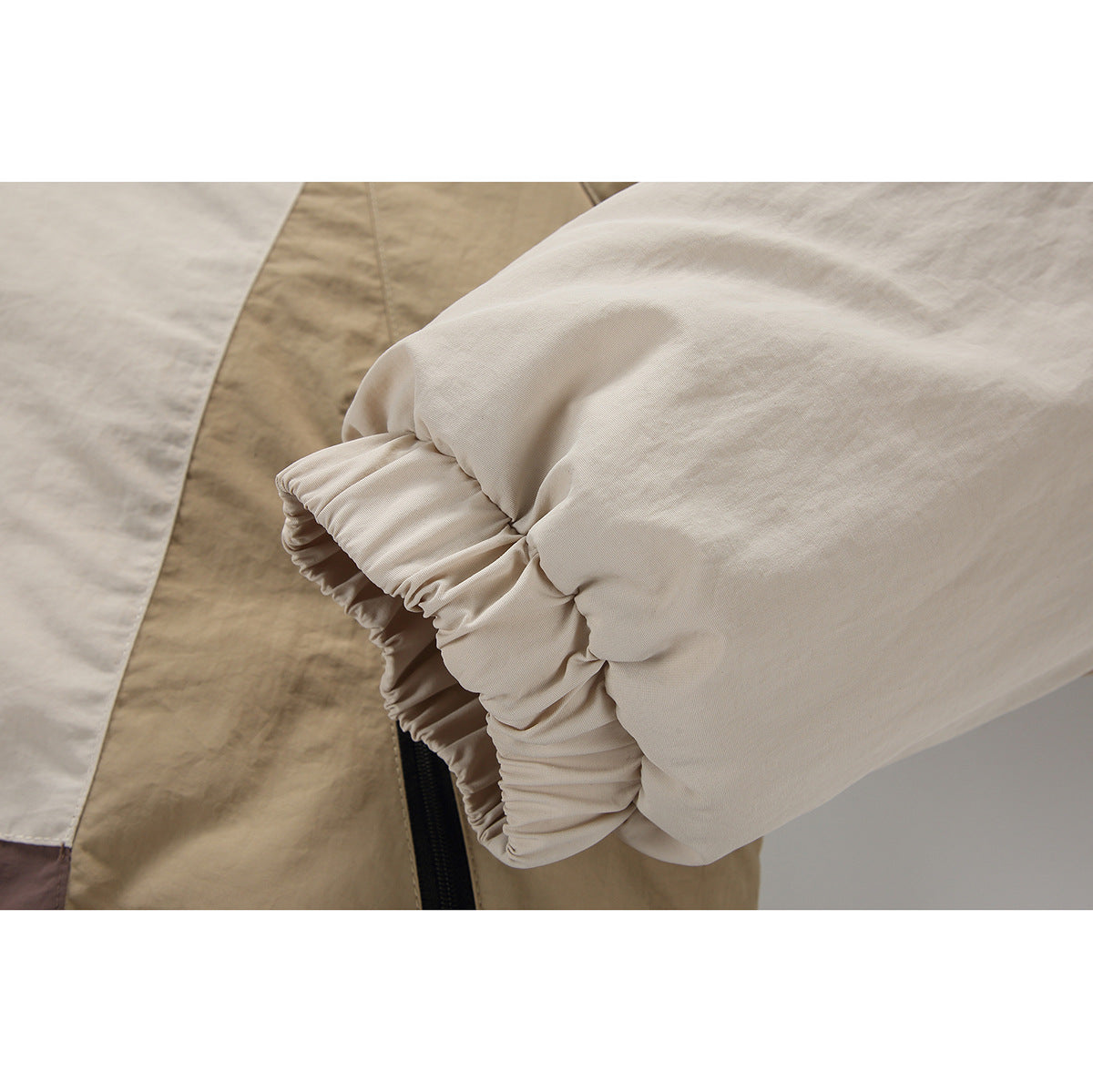 Close-up of a cream jacket sleeve with an elastic cuff, tan color-block panel, and part of a black zipper.