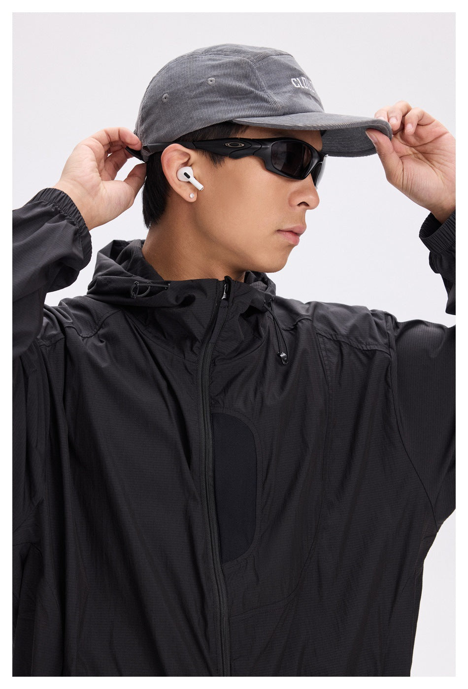 Close-up of a model in a black hooded jacket and gray cap, showing the hood drawcords and chest panel.