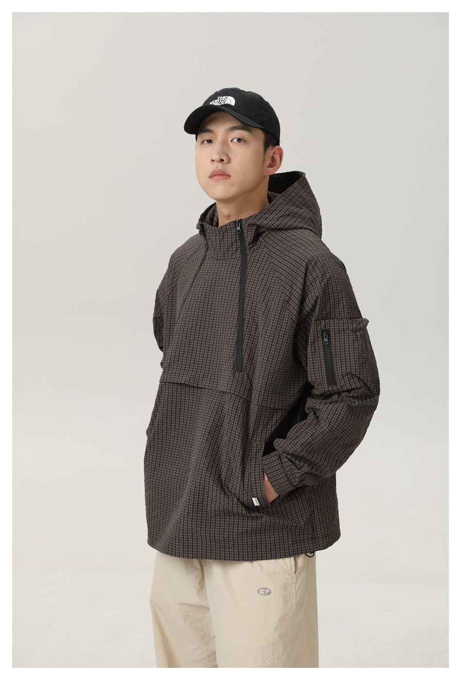 Model wearing a dark brown checked hooded pullover jacket and beige trousers, shown from the thighs up.