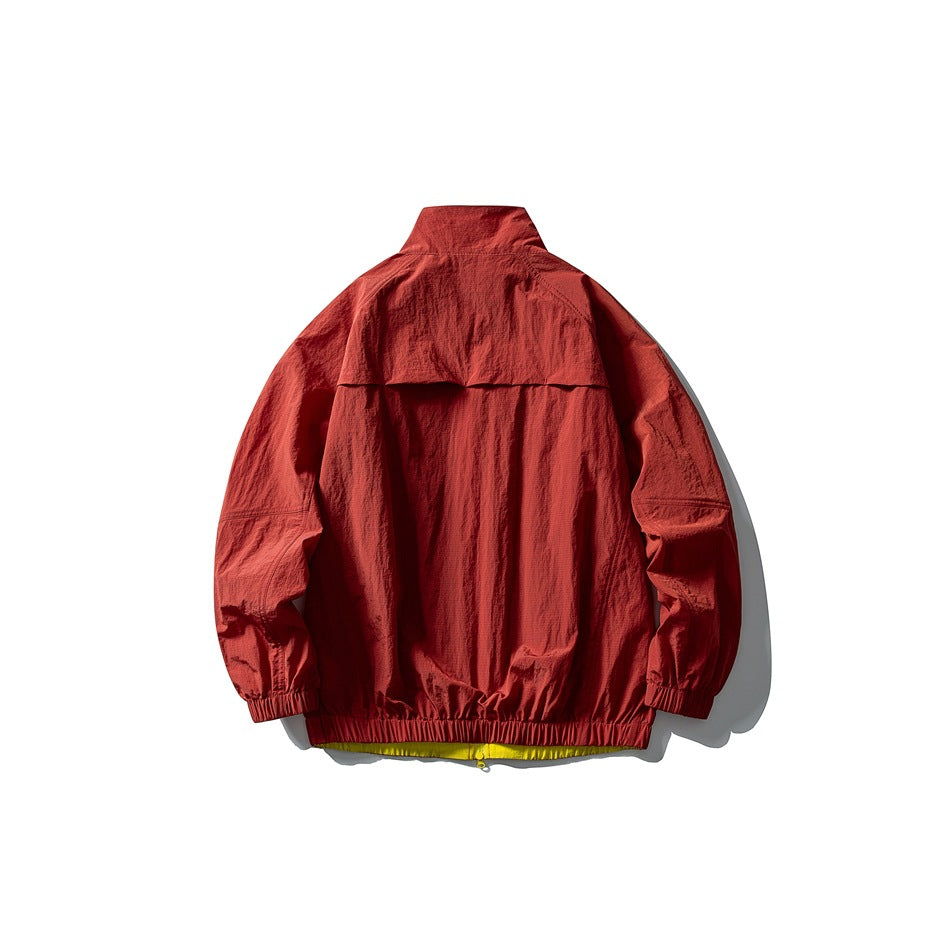 Back view of a rust-red jacket laid flat, with a stand collar, back yoke, elastic cuffs and hem, and a glimpse of yellow