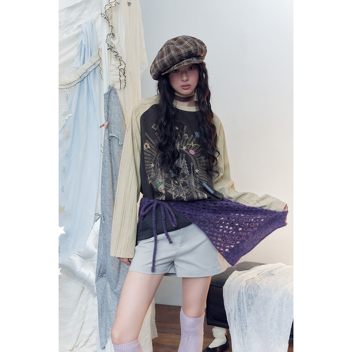 Model wearing a charcoal castle-print raglan top with cream striped sleeves, pale shorts, and a purple knit wrap.