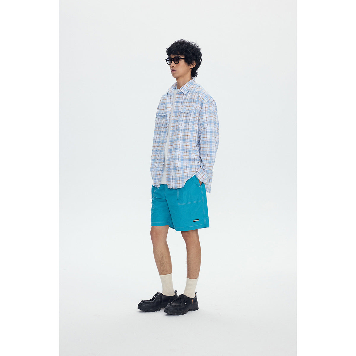 Model wearing a blue-and-tan plaid button-up shirt with chest pockets and turquoise shorts, shown from the front.