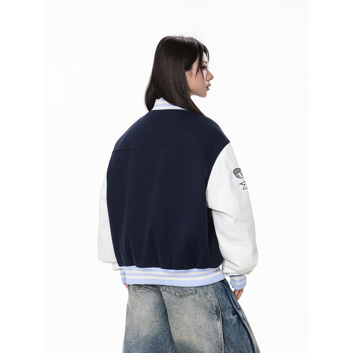 Model turns to show the back of a navy varsity jacket with white sleeves and pale blue striped trim.