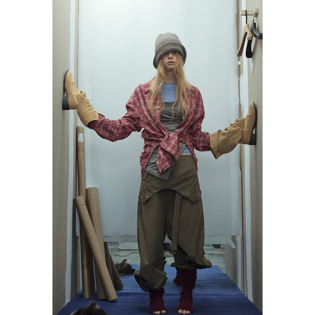 Model wearing a red plaid shirt tied over layered tops, loose olive trousers, a gray beanie and oversized tan mittens.