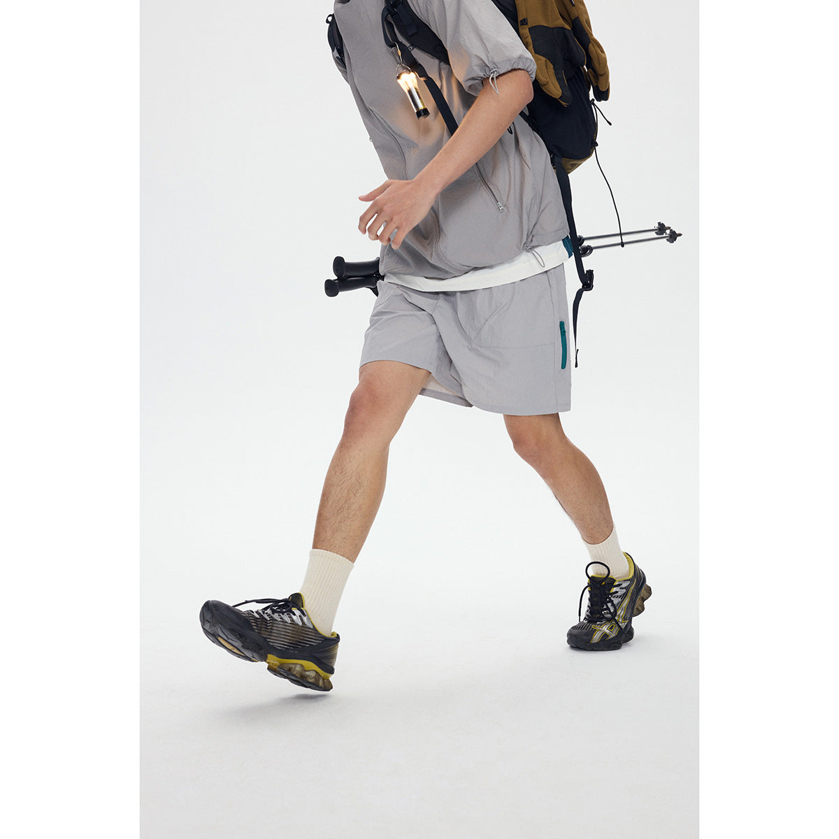 Cropped view of a person walking in a light gray outdoor shirt and shorts with a hiking backpack and trekking poles.