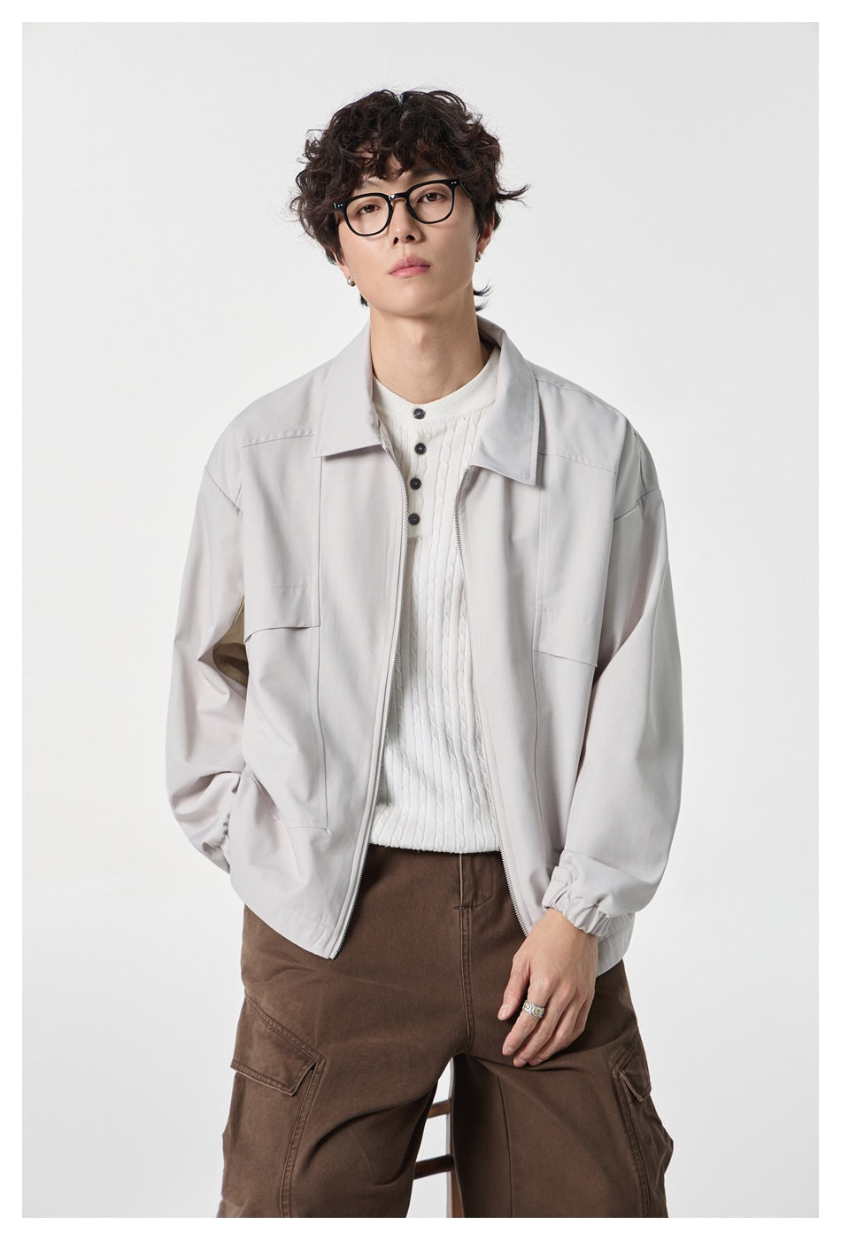 Model seated in an open beige zip jacket over a white cable-knit henley, with brown cargo pants