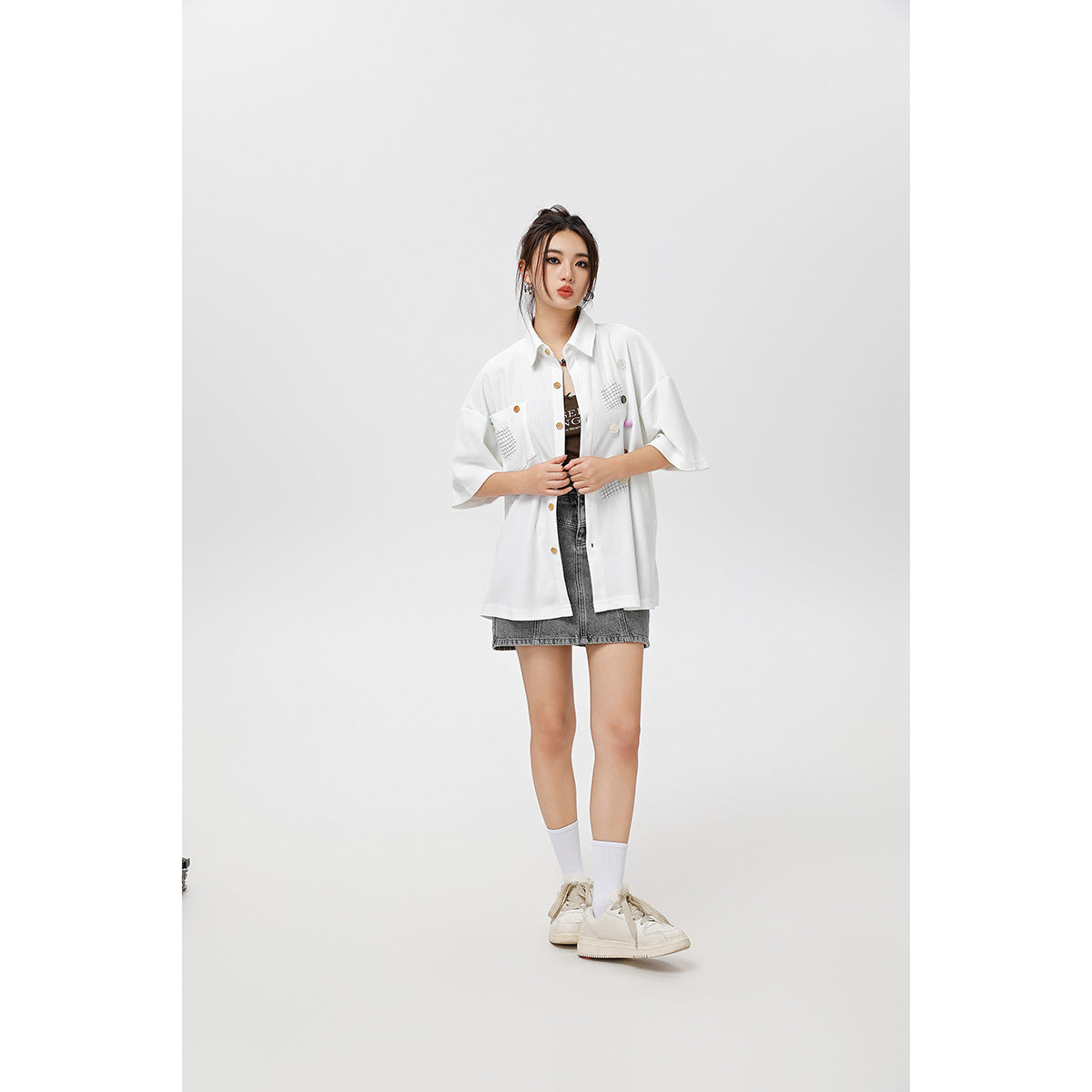 Model wearing an open white short-sleeve button-up shirt with grid patches and colorful details over a dark top.