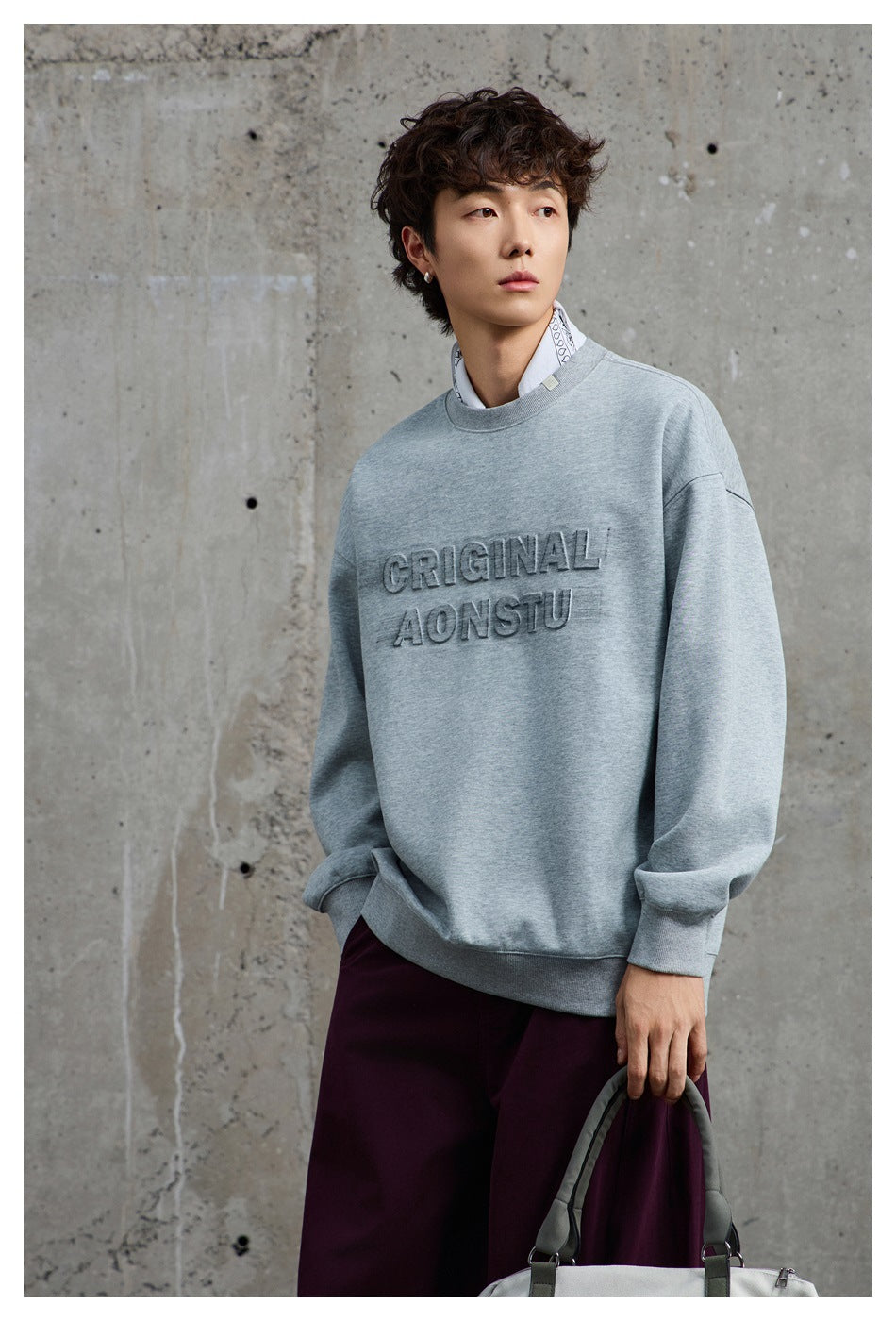 Model in gray embossed-lettering crewneck sweatshirt and burgundy pants, holding a bag against a concrete wall