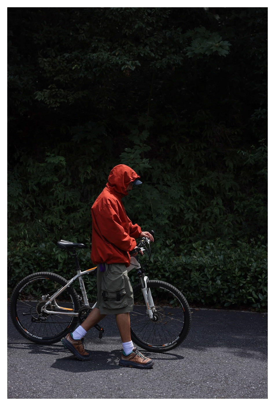 Side view of a person in a red hooded jacket walking a bicycle along a wooded road.