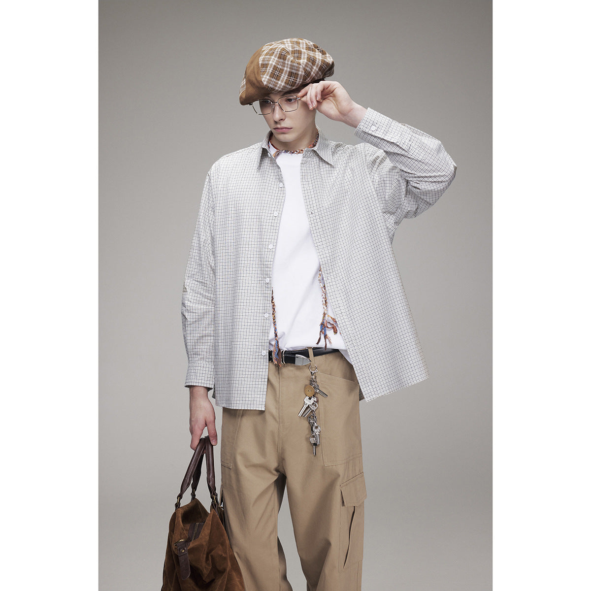 Model wearing an oversized light gray checked button-up shirt over a white tee, with tan cargo trousers and a plaid cap.