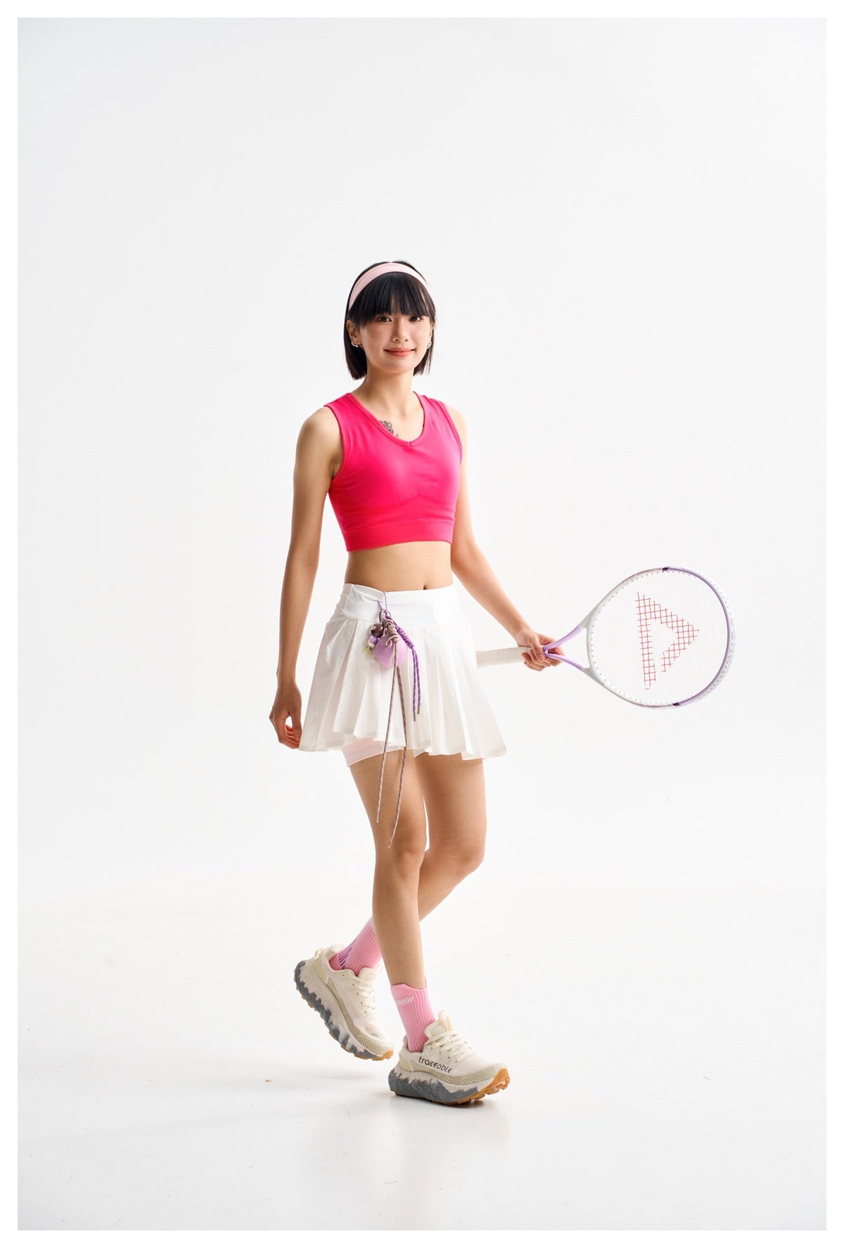 Model in a hot-pink sleeveless crop top and white pleated tennis skirt, holding a racket against a white studio background.
