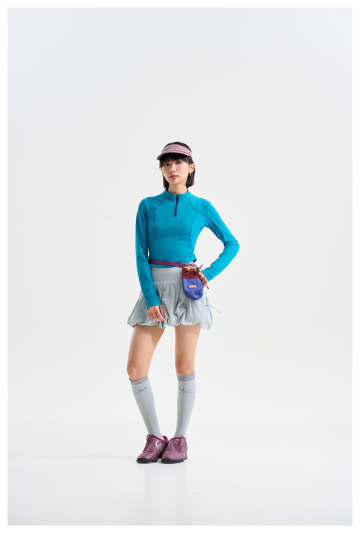 Model in a teal quarter-zip top and pale blue-gray gathered skirt, front view, with a small purple pouch at her waist.