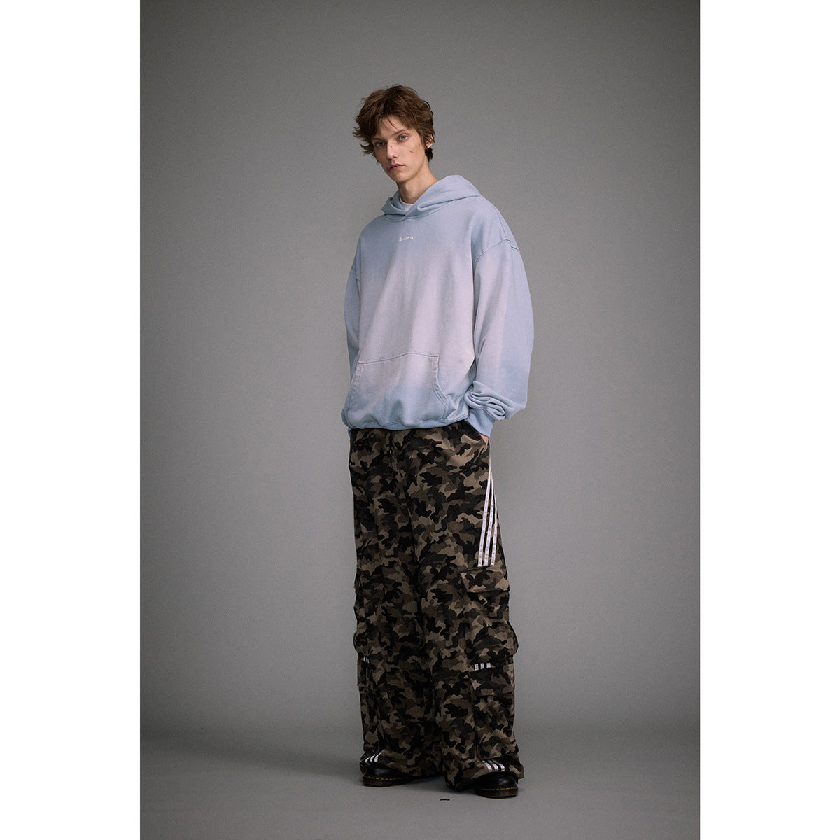 Model wearing a pale blue oversized hoodie with a kangaroo pocket and dark camouflage cargo pants.