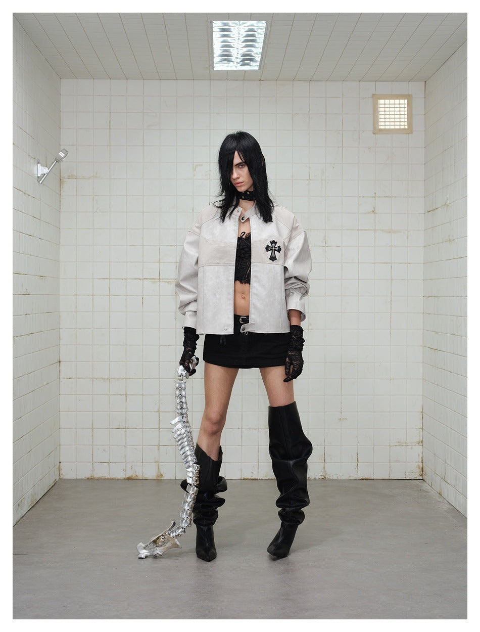 Model wears a pale gray cropped jacket with a dark cross emblem, black mini skirt, lace gloves and tall slouchy boots.