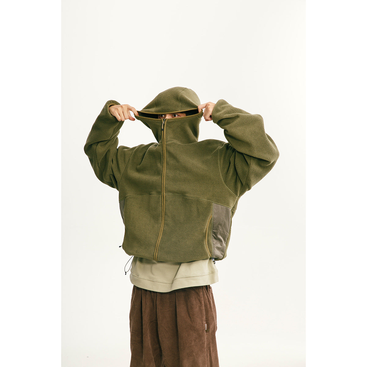 Model wearing an olive hooded zip-up fleece jacket with contrasting side panels, pulling the high collar over the face.