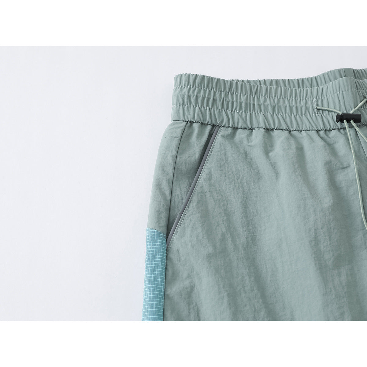 Close-up of the pale sage pants' elastic waistband, drawcord and slanted pocket with a blue grid side panel.