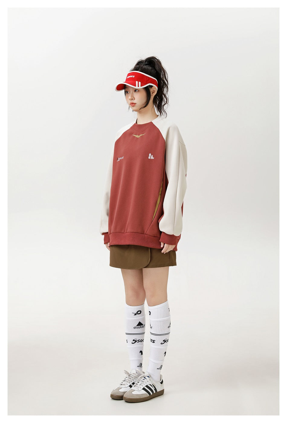 Model wearing a brick red oversized raglan sweatshirt with cream sleeves, brown skirt, red visor and white socks