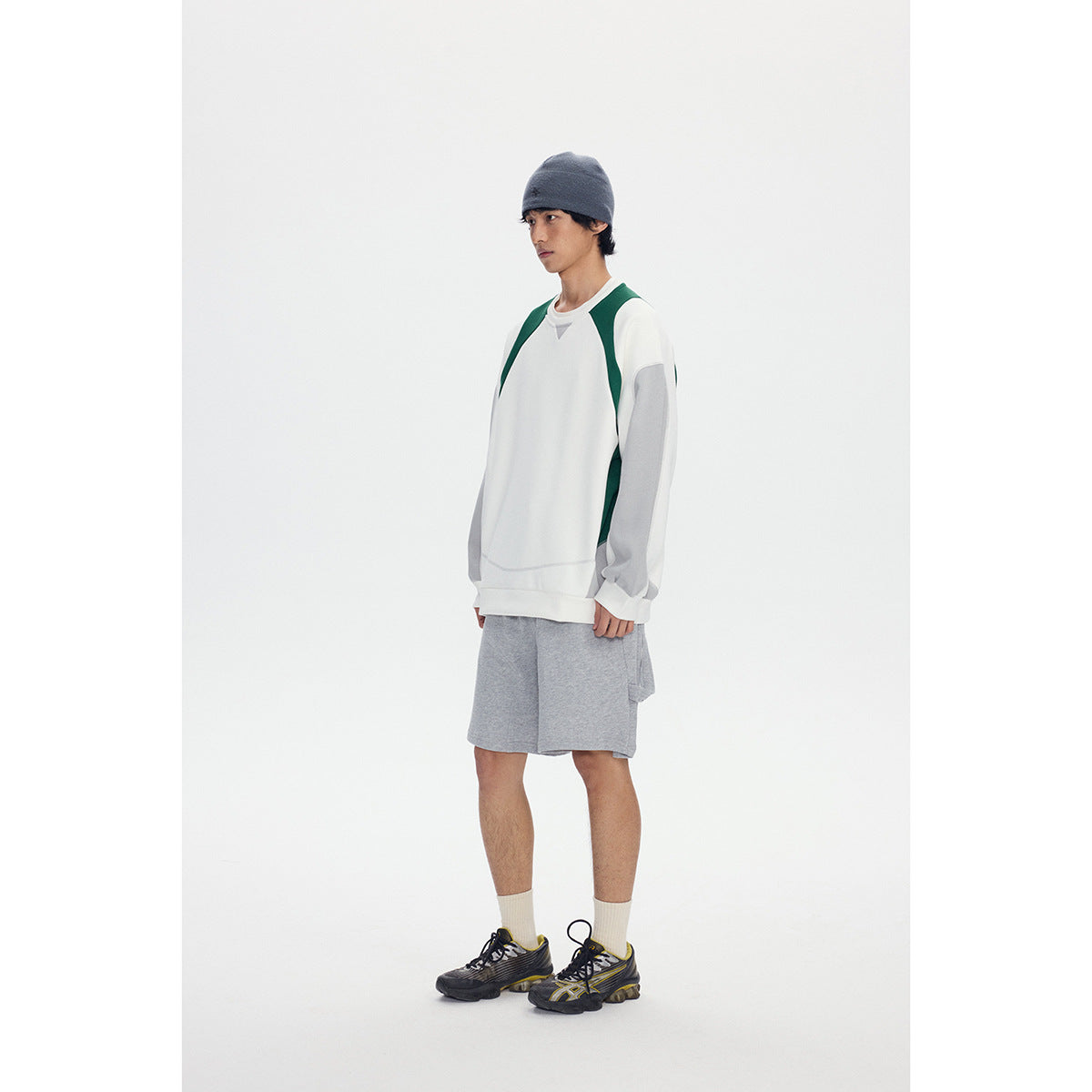 Model wearing a white sweatshirt with green and gray panels and gray shorts, shown standing in a studio.