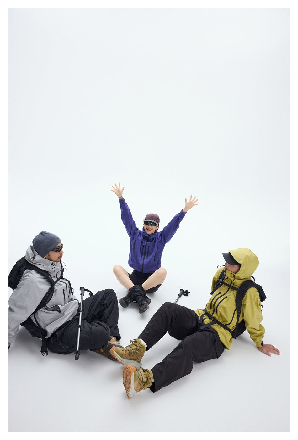Three hikers pose seated in gray, purple, and yellow hooded jackets with black pants and backpacks.