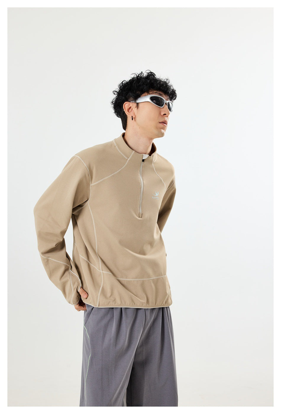 Model wearing a tan long-sleeve quarter-zip top with contrast seams and loose gray pants, shown from the front.