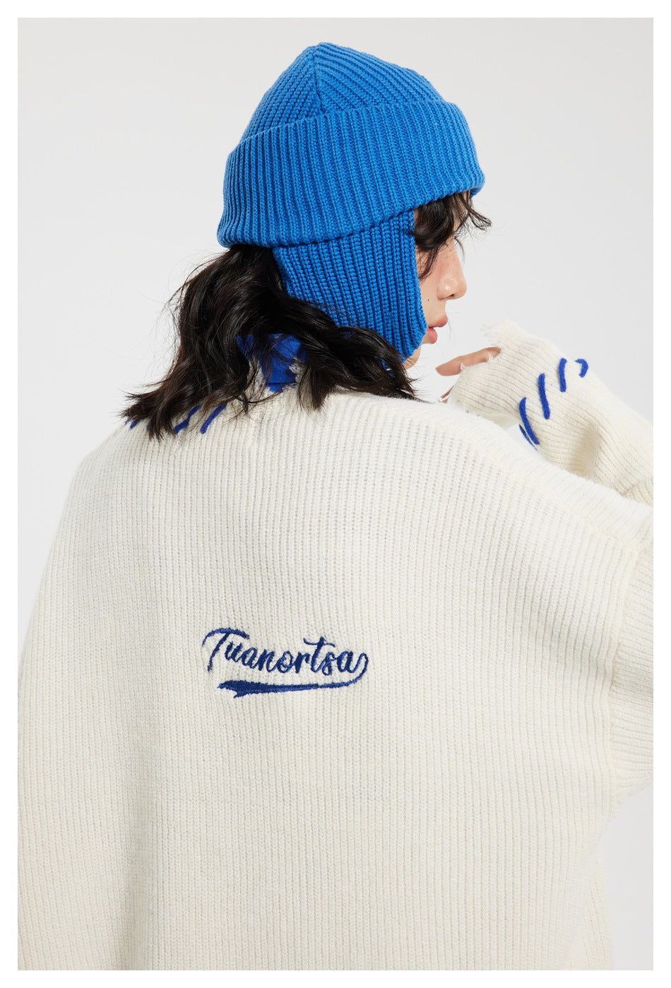 Back view of a model wearing an ivory ribbed sweater with blue embroidery and a bright blue knit earflap hat.