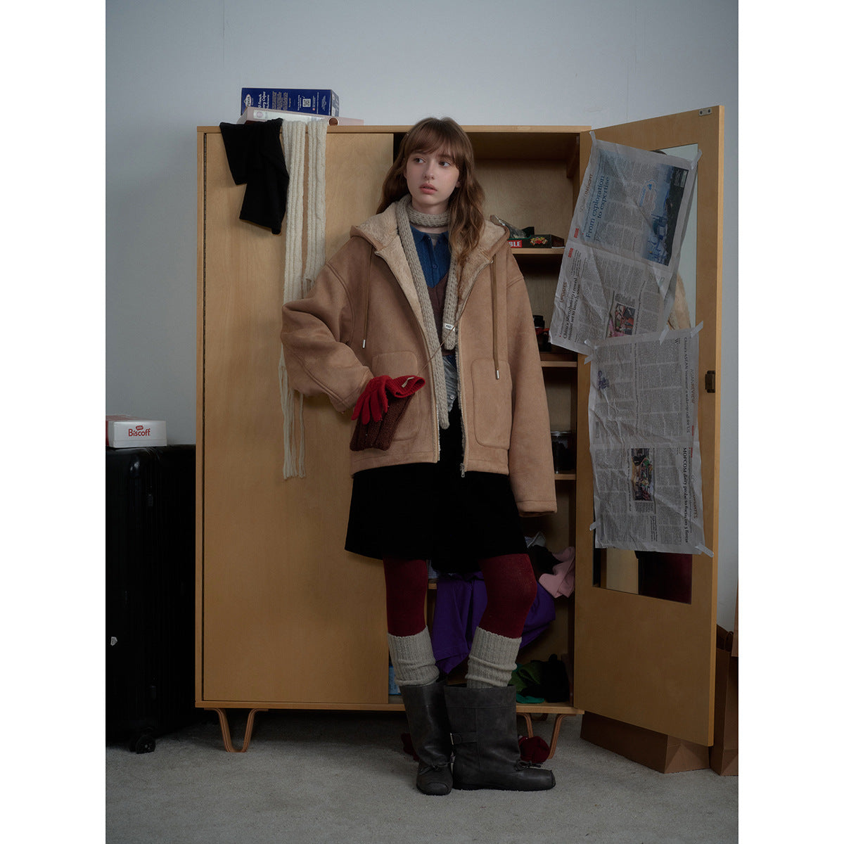 Full-length model wearing an open tan shearling-lined hooded jacket with layered knitwear, a black skirt and burgundy tights.