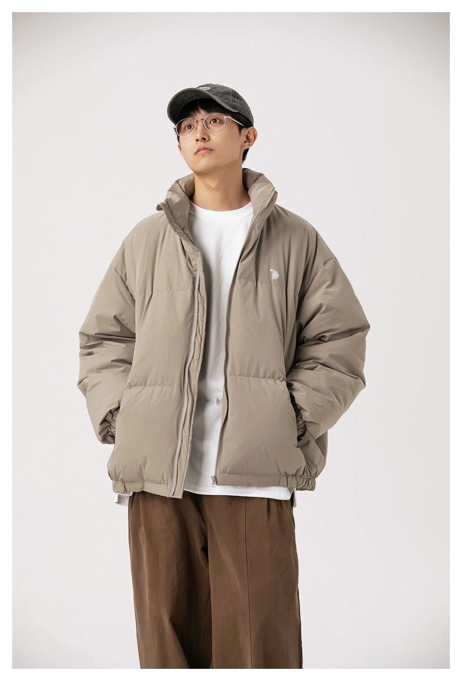 Man wearing a taupe-gray puffer jacket with stand collar, hands in pockets, white tee and brown trousers