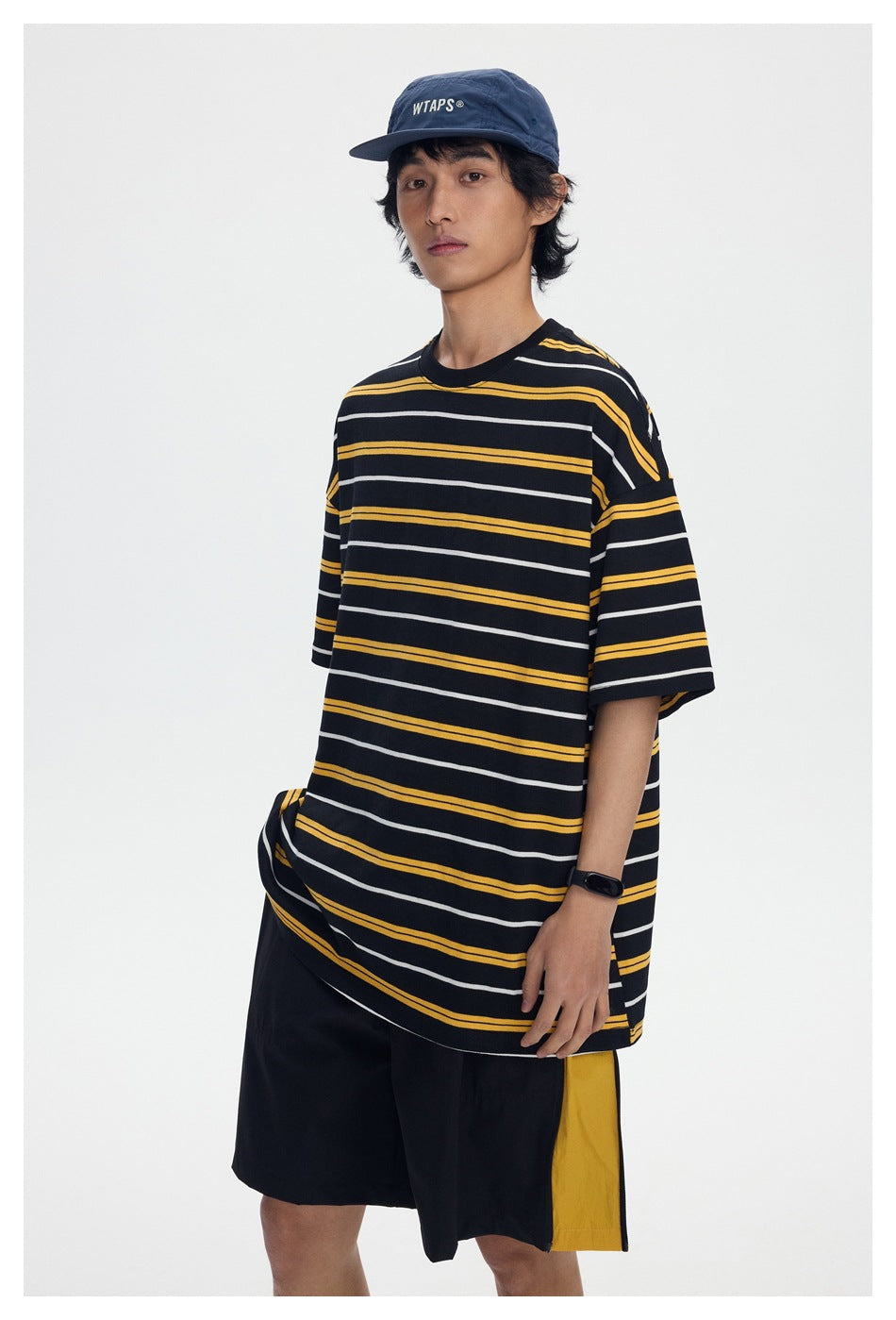 Model wearing a black oversized T-shirt with yellow and white stripes, black shorts, and a navy cap.
