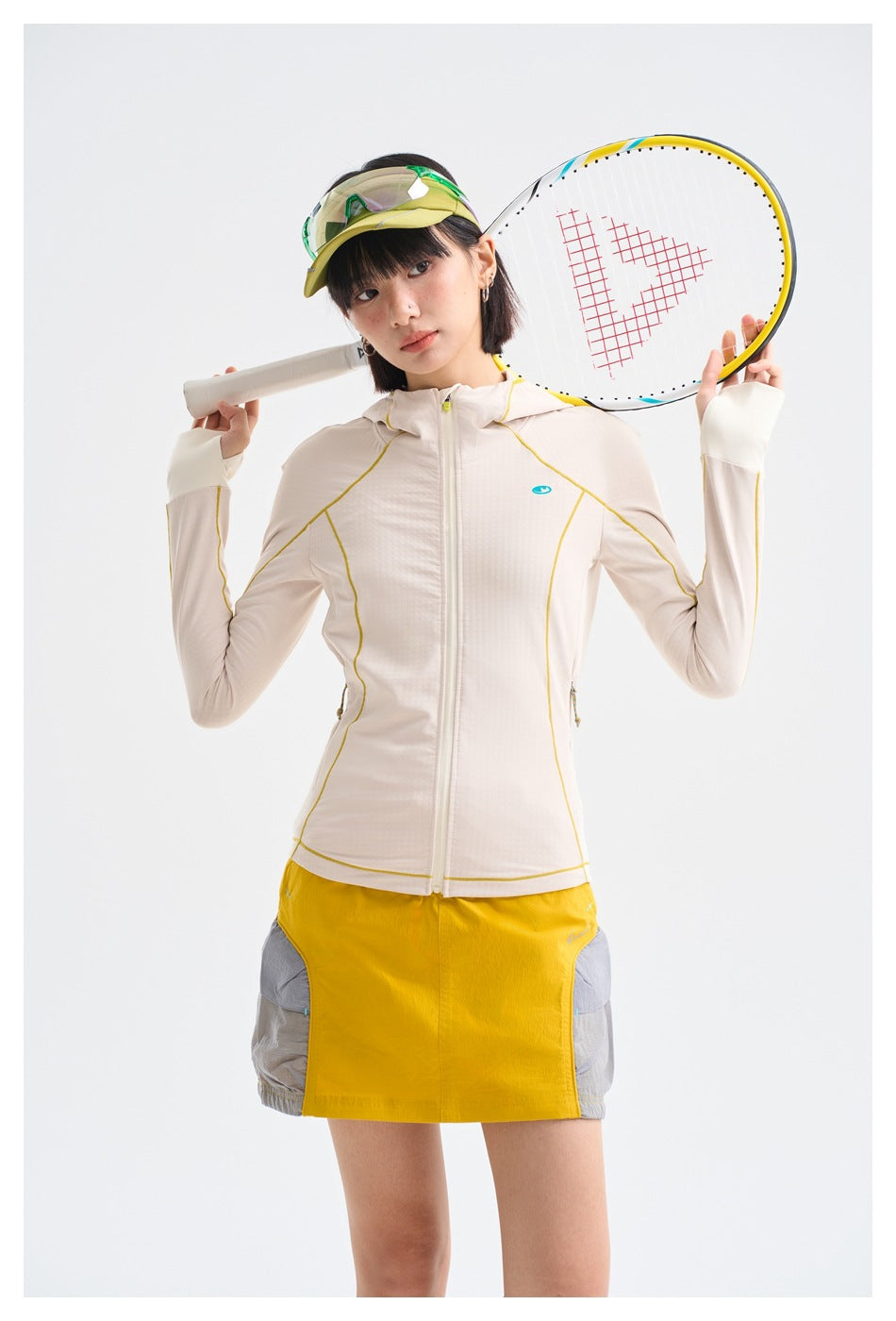 Model wearing an ivory hooded zip jacket with yellow piping and a yellow-and-gray skirt, holding a tennis racket.