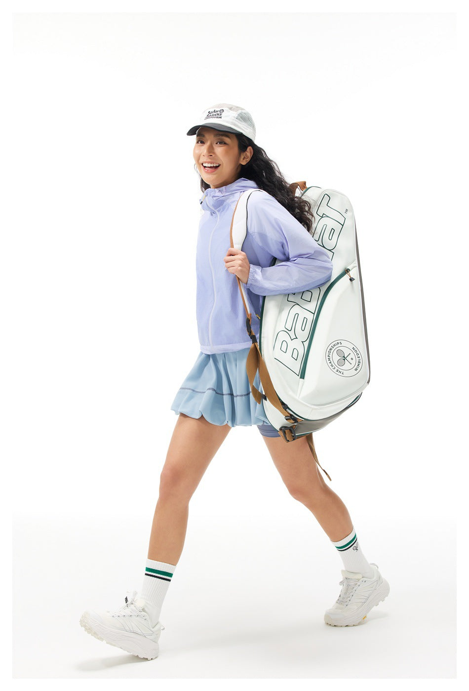 Model in a pale lavender zip jacket and light blue pleated tennis skirt, carrying a large white tennis bag.