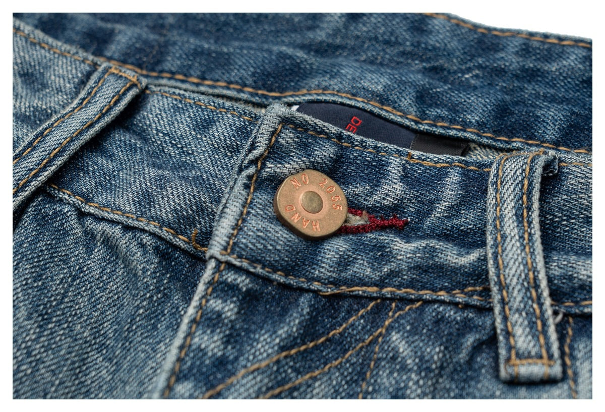 Close-up of blue distressed jeans showing the waistband button, belt loop, contrast stitching and inner label.