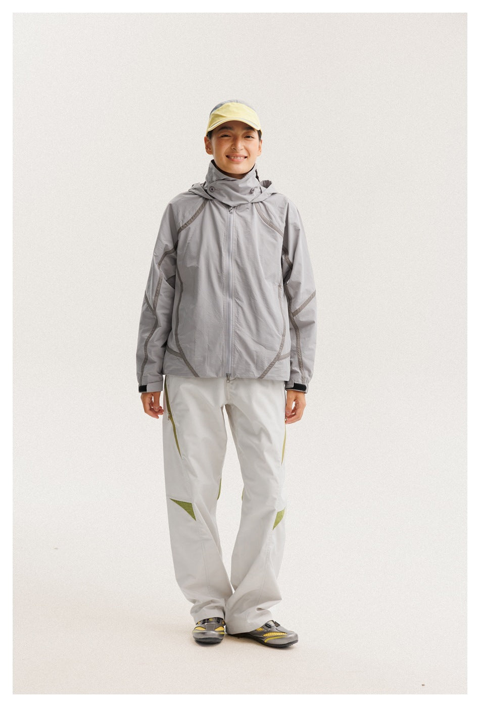 Full-length model wearing a light gray hooded zip jacket with darker seam details and white pants with olive accents.