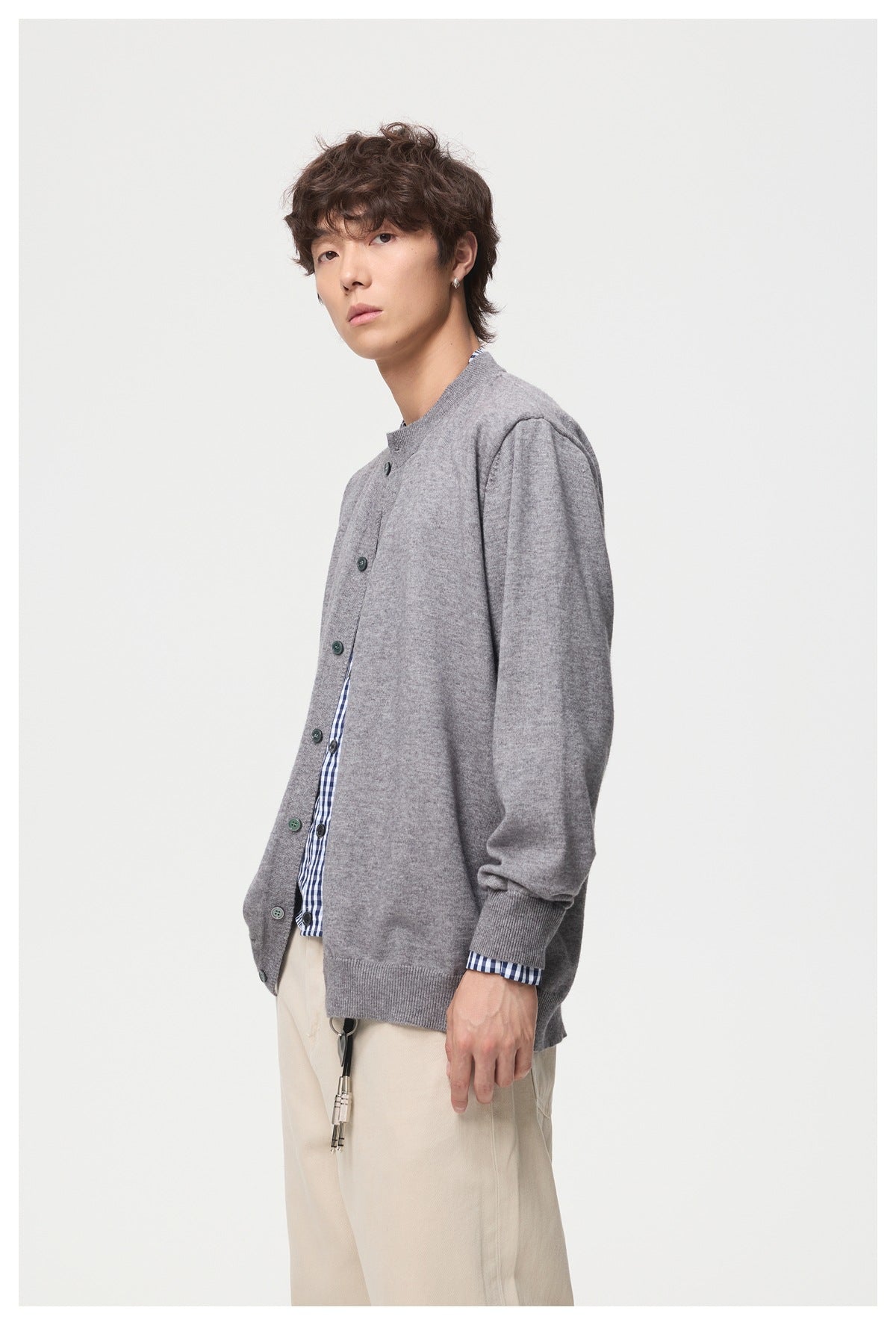 Side-angle view of a model wearing a gray button-front cardigan over a checked shirt and cream trousers.