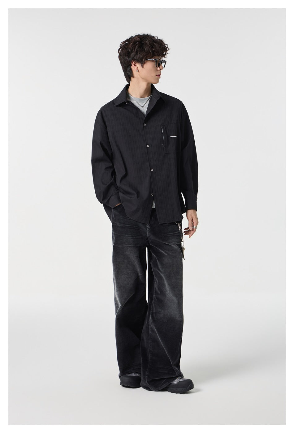 Model wearing a black button-front shirt with a chest pocket, layered over a gray tee and paired with wide black jeans.