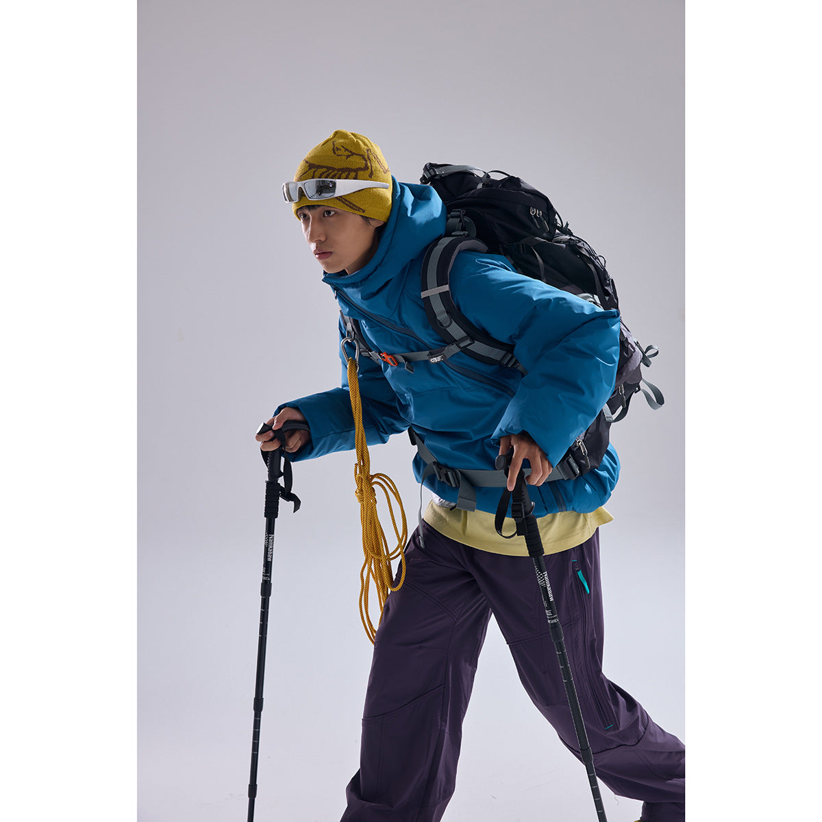 Model wearing a bright blue hooded winter jacket and purple pants with a yellow beanie, backpack, and trekking poles.