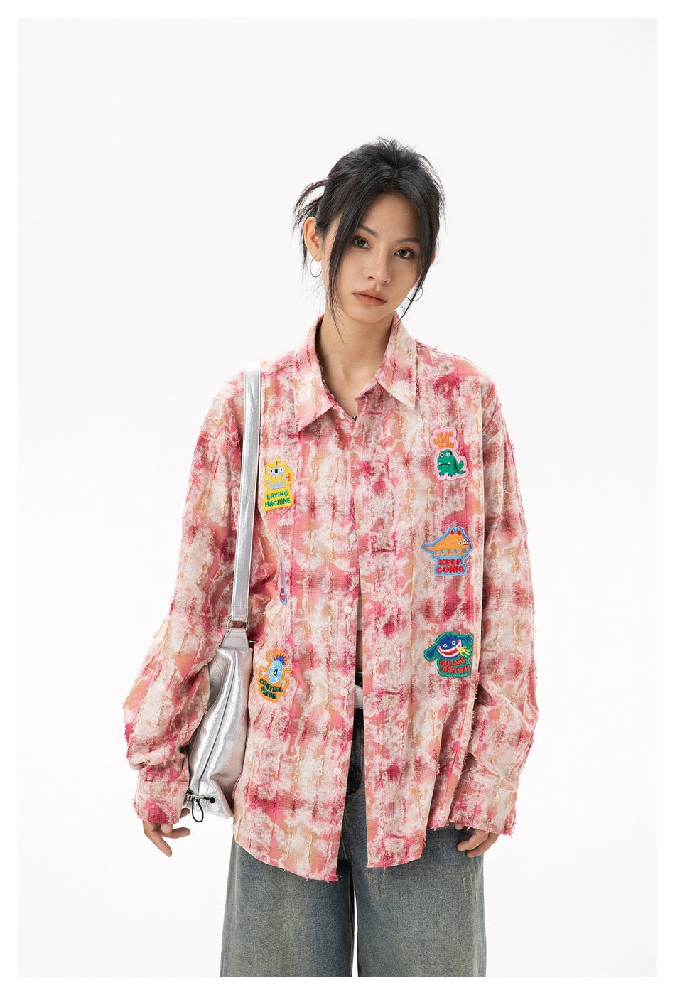Model wearing an oversized pink-and-cream patterned button-up shirt with colorful cartoon patches, blue jeans and a silver