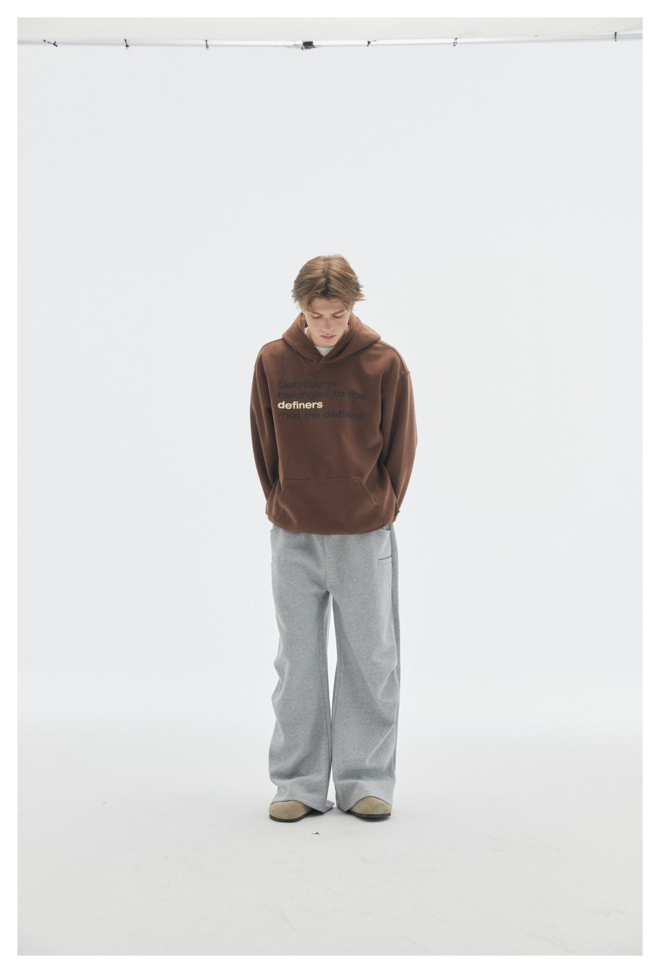Model wearing a brown text-print hoodie and loose gray pants, standing front-facing against a white studio background.