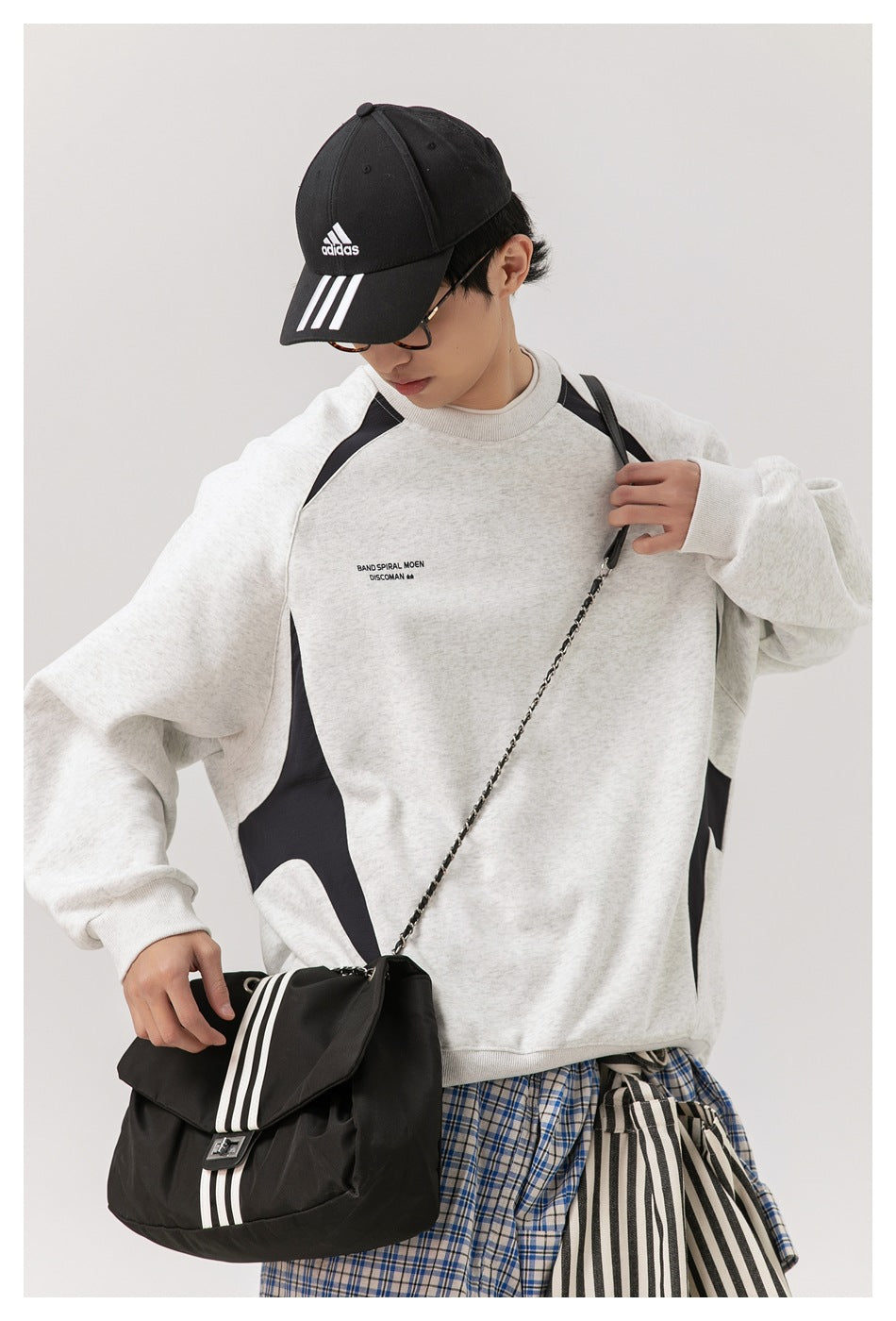 Model wearing a light gray raglan sweatshirt with black side panels and shoulder accents, styled with a black cap and bag.
