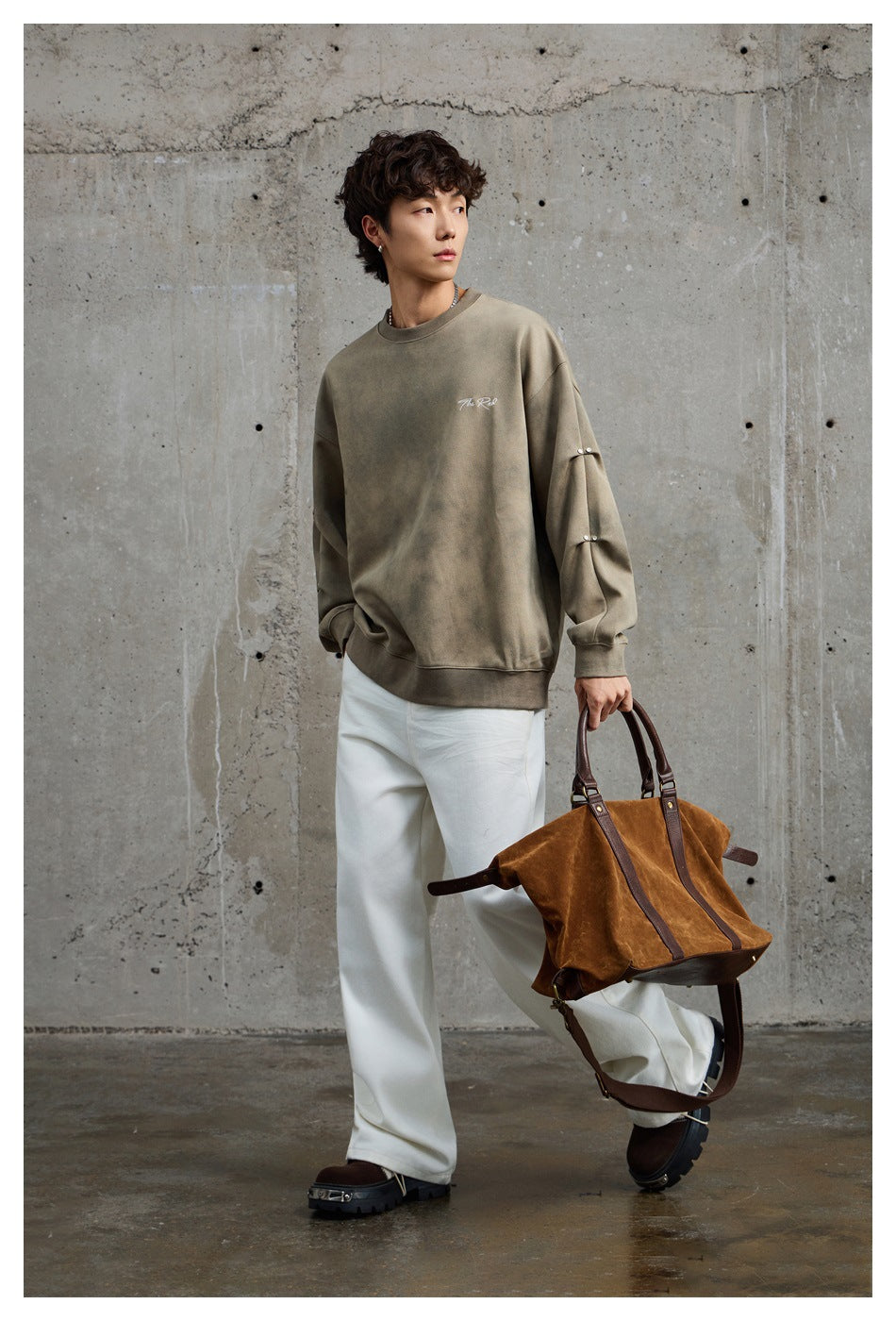 Full-length view of a model in an army green oversized crewneck sweatshirt, white wide pants, and a brown suede bag